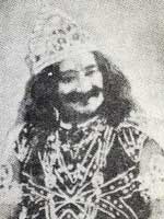 A man with a moustache and long hair dressed with an Indian-style crown along with an elaborately patterned shirt and armbands.