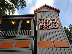 The SooperDooperLooper's station as seen from the ground view. The façade of the station has the roller coaster's name in black text with an orange background on the right tower. The queue building is colored grey with orange fixtures.