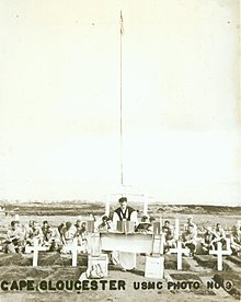 Service personnel with a padre at a memorial service. In the foreground are several graves marked with white crosses.