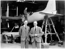 Photograph of two men in suits, in front of an aeroplane, facing the camera.