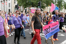 A group of men and women, dressed in purple, pink and blue, marching down a street with a banner which reads "Out and proud – Bisexuals! – Fighting for equality"