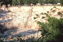 Gathering of people at top edge of sinkhole appear tiny compared to sloping sides of sinkhole about 70 feet deep and a few hundred feet across. Debris is scattered on slope and floating in dirty water in bottom of sinkhole.