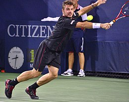 A man wearing a black shirt, black shorts, and black shoes staggers to his left to return a tennis ball as he holds his racket out on the right hand in front of his body