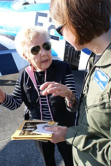 Olsen as an elderly woman speaking with a younger woman dressed in a flight suit, in front of a P-51 on display.