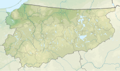 Mapa konturowa województwa warmińsko-mazurskiego, po prawej znajduje się owalna plamka nieco zaostrzona i wystająca na lewo w swoim dolnym rogu z opisem „Roś”