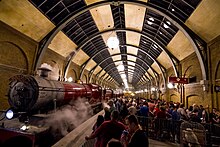 On the left is a red steam locomotive and three carriages, with steam drifting out above. On the right passengers queue behind barriers and signs that say "Hogwarts Express" and "Platform 9+3⁄4". Over both of these is a large cylindrical glass and steel roof.