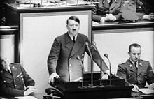 Picture of a man in a suit with a mustache who looks like Hitler speaking behind a microphone.