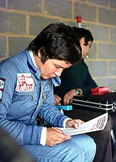 A woman in racing overalls is holding paper in both her hands and reading the material printed on it