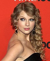 Taylor Swift se encuentra en un área de prensa de Time. Tiene el pelo rizado y lleva un vestido negro sin tirantes.