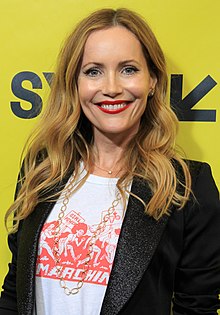 Leslie Mann wearing a white print t-shirt and black blazer, grinning at camera, in front of a bright yellow backdrop