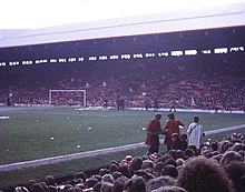 Photograph of a stand full of people. In front of the stand is a field and a goal.
