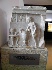 White relief depicting a youth ladlind wine from a krater, a type of container, next to a round table with vases.