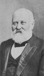 portrait shot of a man in late middle age with white hair and beard. He is seen to wear a suit with vest and a small bow-style tie.