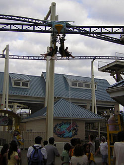 Pictured is one of the Roller Soaker cars traversing the suspended track's layout. Below people observe the track hanging overhead, with a water geyser spraying water at the ride's car. The roller coaster's station is featured in the background.