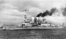A large warship, thick black smoke pouring out of its rear funnel, steams through the calm sea. The ship's forward funnel has been merged via a diagonal section into the one behind it, an a larger mast with an observation platform has been installed in place of the original mast.