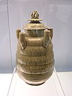 A tan jar with several rounded bands carved into the body and five upward-facing tubes equidistantly spaced along the top area of the jar. The lid is crowned with a lotus flower carving.