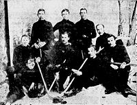 Men wearing hockey sweaters assembled in three rows in front of a painting of a forest scene. Several are holding ice hockey sticks