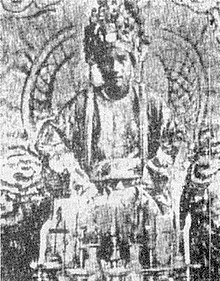 A black and white photograph of a young cleanshaven man wearing an elaborate headdress and sitting on a throne. He is wearing robes and in front of him are some accoutrements on a table.