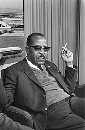 A middle-aged black man in a suit, seated in an airport lounge with a cigarette