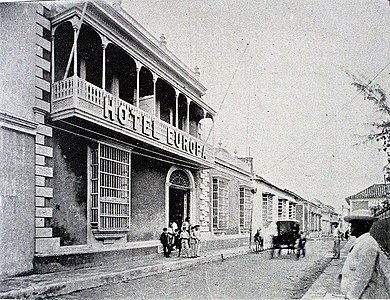 Black and white photograph showing the Hotel Europa in Maracaibo around the time the film was shot. The hotel is on the left with a group of people standing in front of it. A horse and carriage are seen on the road in the middle of the picture, and several men are standing on the right.