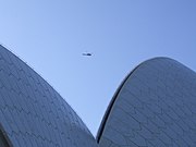 Vrh neprepoznavne valovite stavbe pod modrim nebom in helikopter, ki je tako oddaljen, da je podoben vrabcu