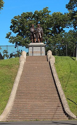 Photograph showing a long, broad flight of steps leading to a large metal statue of two men. There are about 50 steps. The two men in the statue are roughly life-sized, and are standing on a large stone pedestal. The pedestal has bronze plaque that reads "Goethe und Schiller".
