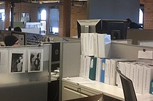 Thick white binders in a cubicle, three people are visible, exposed brick walls