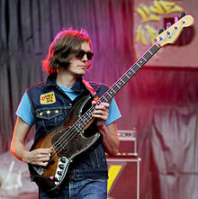 Fraiture plays an electric bass on stage, wearing sunglasses and a denim vest with a patch saying "Have a nice day".