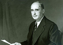 A black-and-white photo, tinted copper green, of a balding man in a tie, suit, and spectacles sitting head turned towards the camera and holding a sheet of paper
