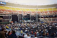 An elaborate concert stage, seen during the day inside a mostly empty stadium. The stage comprises several dark, rectangular structures. Fans are scattered throughout the floor seats, while the stadium seating is empty.