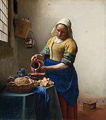 Woman in yellow shirt and blue skirt pouring milk from a pitcher into a bowl on the table beside her