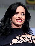 A standing dark-haired white woman, wearing a black top facing towards but looking towards the right of the camera, smiling.