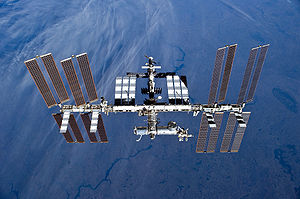 A planform view of the ISS backdropped by the limb of the Earth. In view are the station's four large, gold-coloured solar array wings, two on either side of the station, mounted to a central truss structure. Further along the truss are six large, white radiators, three next to each pair of arrays. In between the solar arrays and radiators is a cluster of pressurised modules arranged in an elongated T shape, also attached to the truss. A set of blue solar arrays are mounted to the module at the aft end of the cluster.
