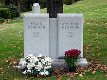 A joint grave marker, though made of multiple pieces of stone if a visible line is an indication. The left and right halves of the marker jut out, giving the impression of individual tombstones while still attached in the middle. The left marker reads "Frank (linebreak) O'Connor (linebreak) 1897 (linebreak) 1979". The right marker reads "Ayn Rand (linebreak) O'Connor (linebreak) 1905 (linebreak) 1982". An arrangement of white flowers sits in front of Frank O'Connor's marker. A pot of red flowers sits in front of Rand's marker. Green grass and dead leaves surround the grave marker. There is a tree immediately behind the marker, but because of the closeness of the camera shot, only the trunk is visible and no leaves can be seen.