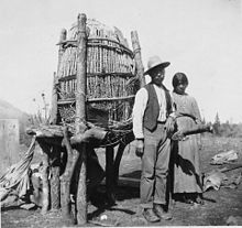 A man and woman of the Mono tribe stand in front of an acorn cache, similar to a large woven basket held up by thick wooden sticks