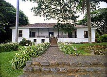 Color photo of a house with white walls, a paved path and stairs leading to a covered terrace at the entrance.