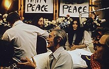Lennon and Ono sit in front of flowers and placards bearing the word "peace." Lennon is only partly visible, and he holds an acoustic guitar. Ono wears a white dress, and there is a hanging microphone in front of her. In the foreground of the image are three men, one of them a guitarist facing away, and a woman.