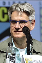 A man with white hair, wearing circular glasses, a blue shirt, and a light brown jacket, faces forward with a water bottle and microphone in front of him.