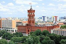 Frontale Farbfotografie vom Roten Rathaus mit seinen roten Backsteinen und Rundbogenfenstern. Das dreigeschossige lange Gebäude wird in der Mitte von einem hohen Glockenturm überragt, der an seinen Ecken mit Säulen verziert ist.