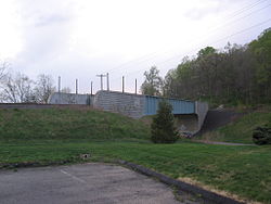 A railroad bridge with one track crossing a road. The bridge is made of blue girders, with stone block abutments. The seal of the state of Connecticut is visible on the closer abutment.