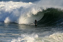 No meio do mar, uma onda está quebrando. No centro da imagem, bem pequeno, um surfista aparece logo antes da arrebentação da onda, sendo que parte está sobre ele.
