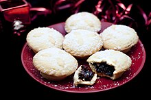 Six mince pies on a plate, sprinkled with sugar and with one broken open