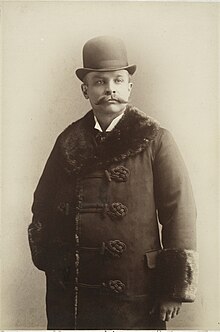 Hip height portrait of middle-aged man with moustache wearing bowler hat and fur-trimmed overcoat
