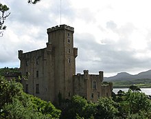 A grey castle with tall square towers stands amongst trees in full leaf.