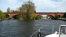Küçük bir botun önünden bakılan, derenin üzerinden geçen, kırmızı tuğlalardan yapılmış, sığ kemerli bir köprü