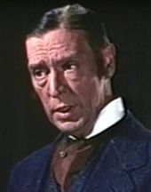 A dark-haired white man wearing a blue suite with a maroon cravat and a white collar