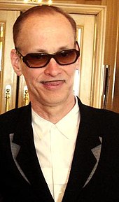 A balding man with a small mustache, wearing sunglasses and a dark suit.