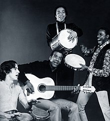 A black and white photo of a man with an acoustic guitar, surrounded by three other men holding percussion instruments