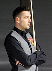 Mark Selby standing with arms folded and holding his snooker cue upright against his chest, wearing black shirt and light grey waistcoat