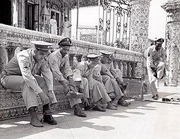 A line of six black American soldiers in service uniform (non-combat) sitting or standing beside the railing at the entrance of a temple. All are taking off their shoes prior to entering the temple.